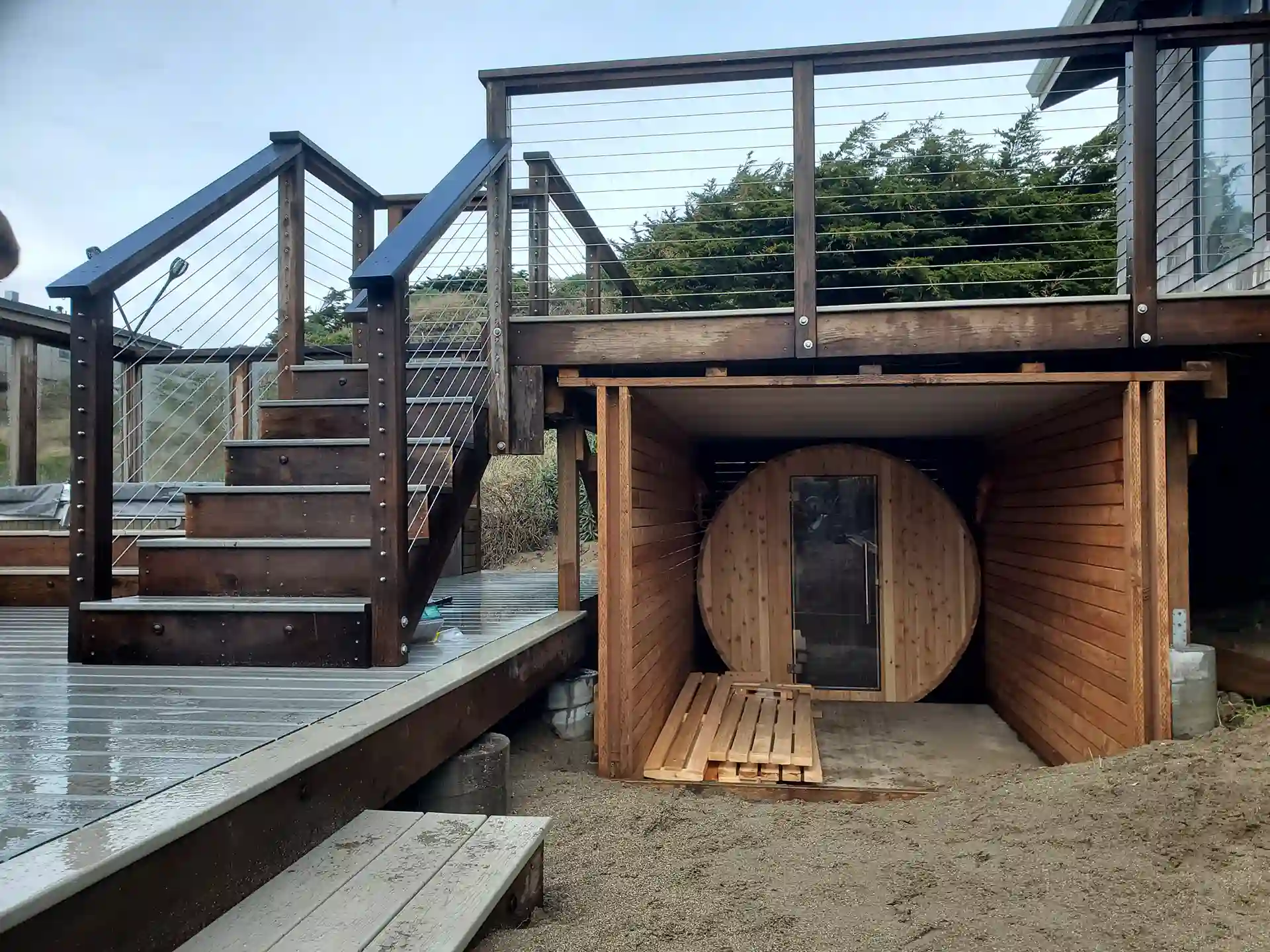 Wood deck with cable railing and stairs, featuring a barrel sauna tucked beneath the elevated structure.