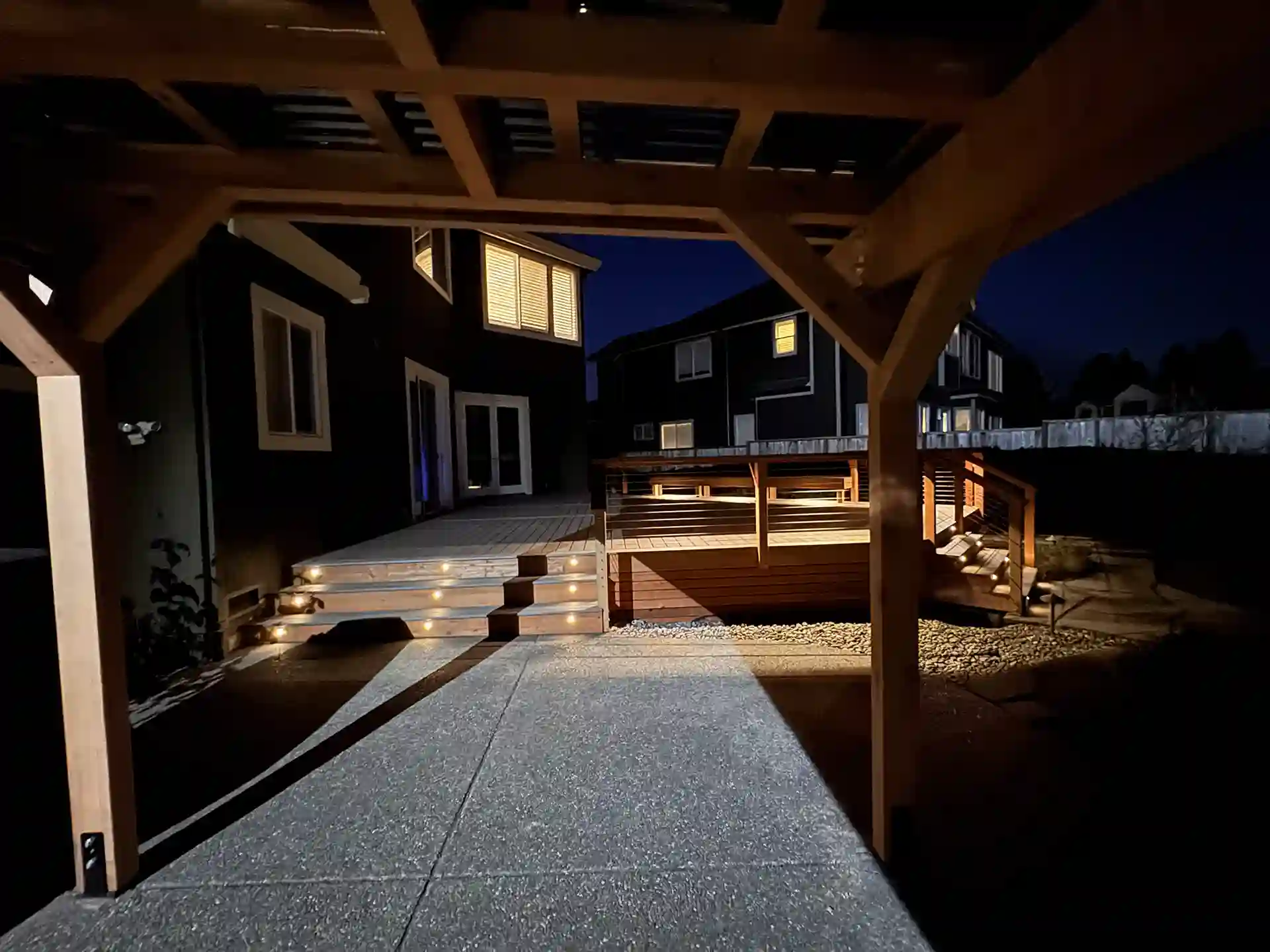 Under-deck view of pergola at night with illuminated deck stairs and railing lighting in a backyard setting