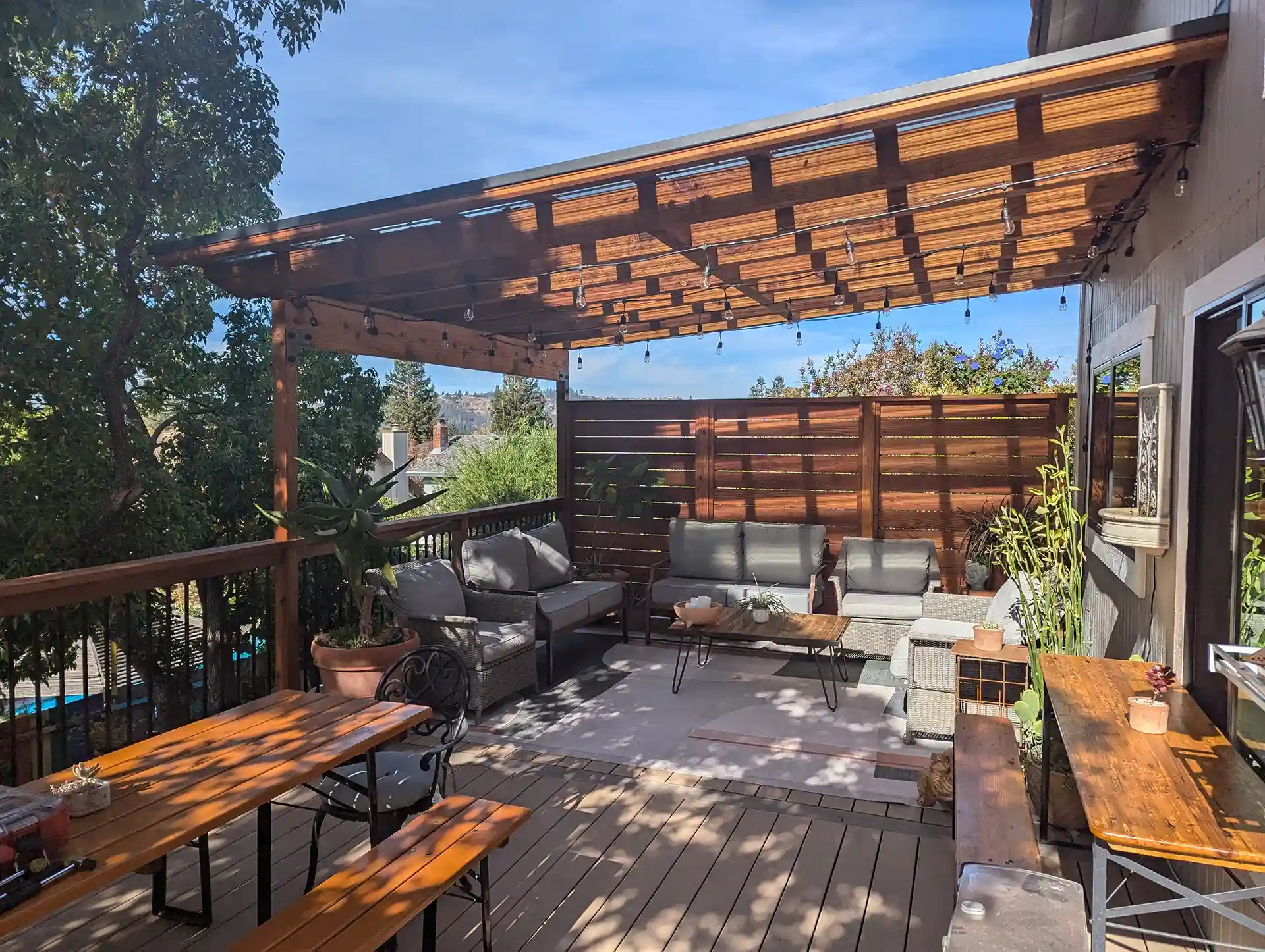 Wooden deck with pergola overhead, cushioned seating, coffee table, potted plants, and string lights forming an outdoor living area.
