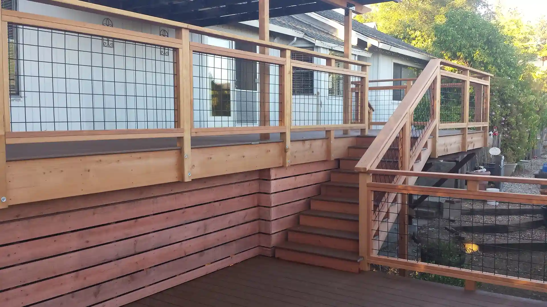 Multi-level wood deck with horizontal skirting, wire panel railings, and central staircase connecting upper and lower levels beside a modern home. 