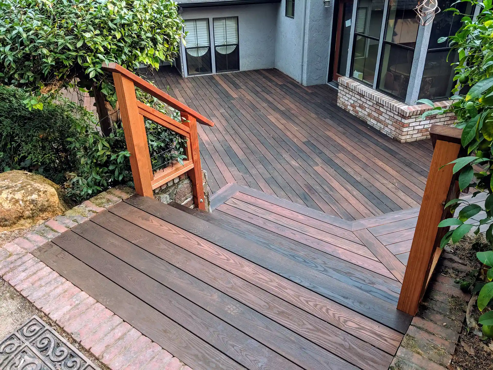 Multi-level wood deck with angled stairs, decorative railing, and rich-toned planks leading to a spacious lower patio area near a brick outdoor feature. 