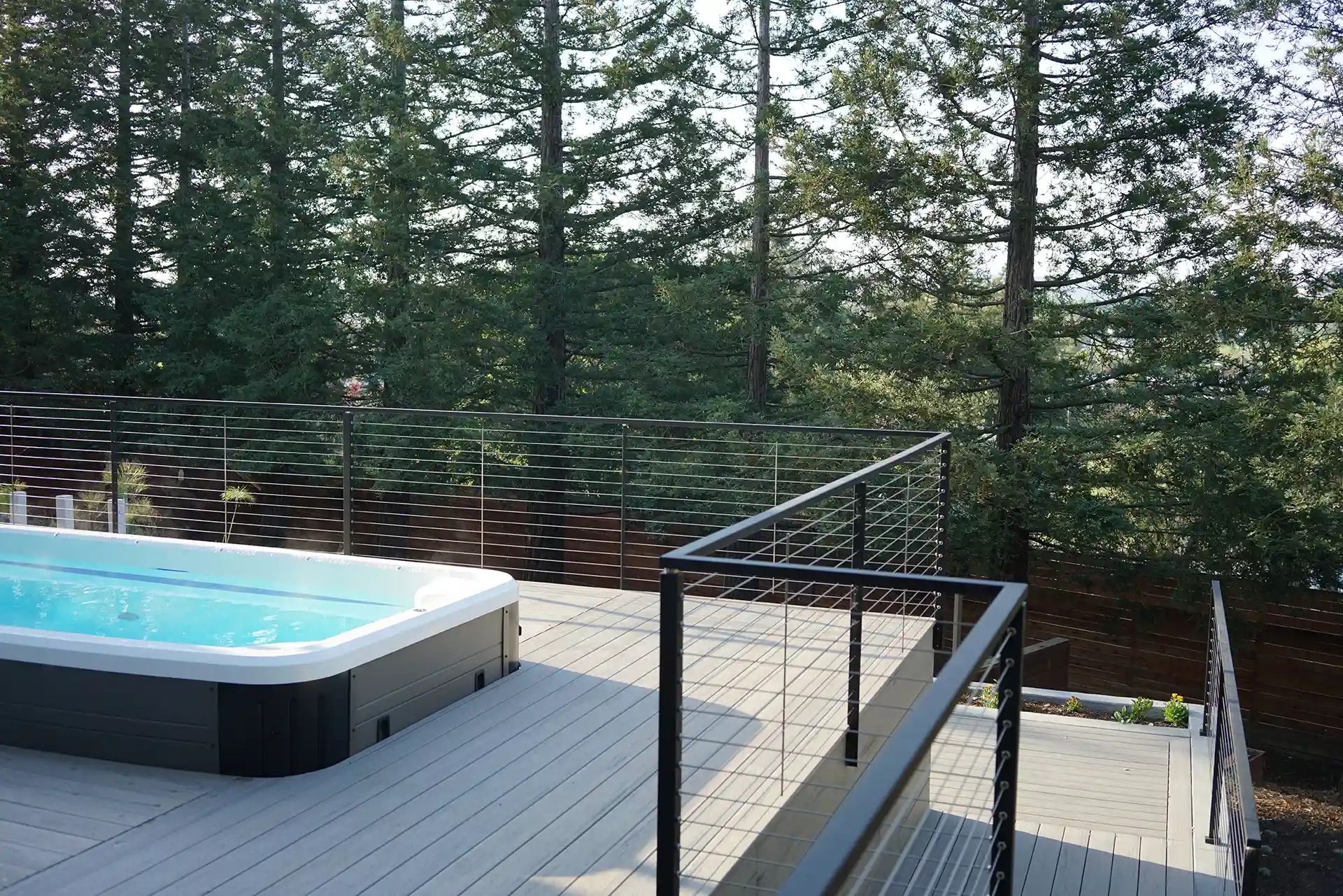 Modern composite deck with gray boards and a metal railing, featuring a hot tub and surrounded by trees.
