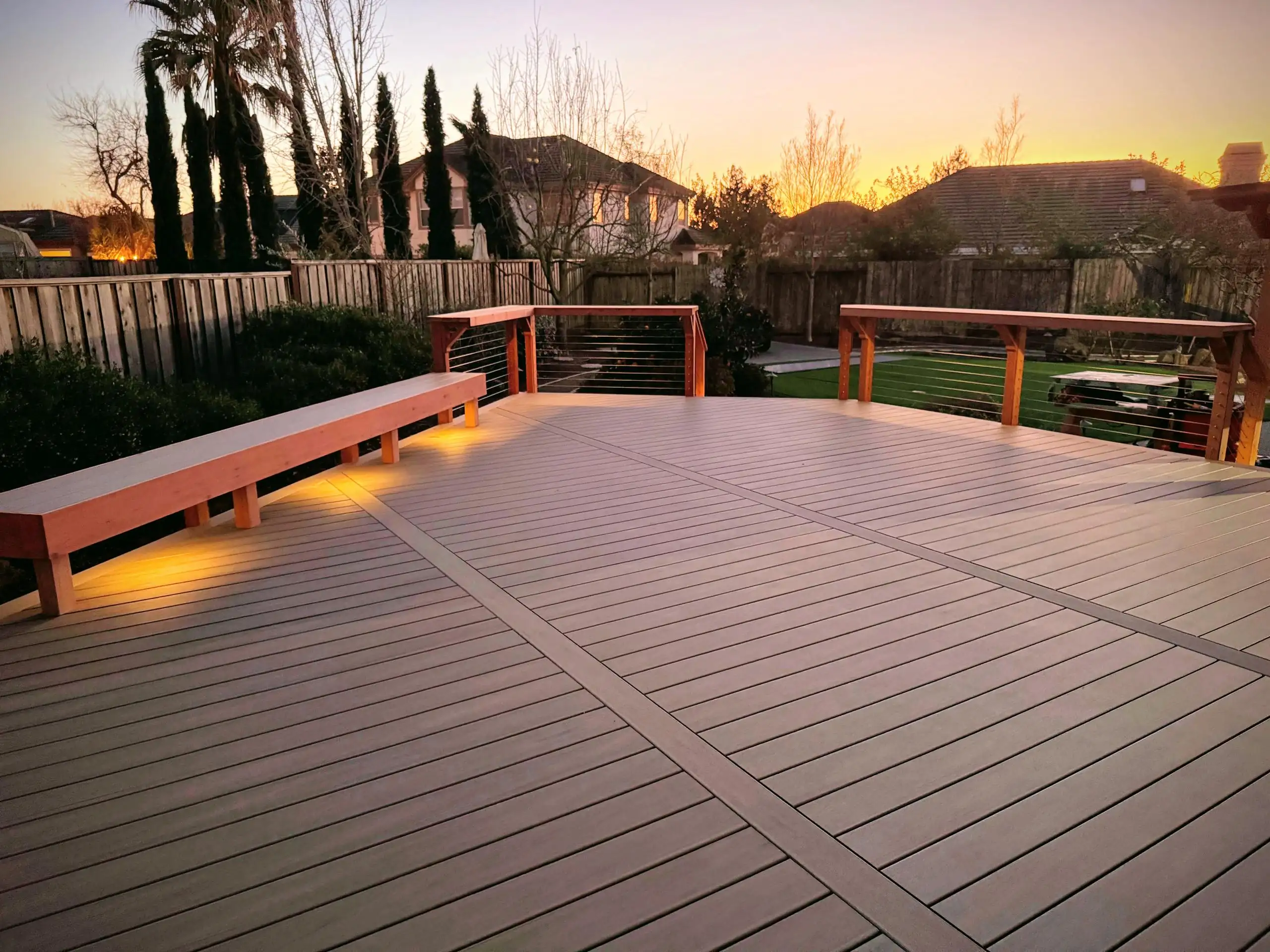 Deck Picture Frame or Not A large, outdoor composite deck with gray, wide-planked flooring, viewed at sunset. The deck features a built-in bench on the left, with hidden lighting underneath. Two sections of railing are visible, each having a wooden top cap/drink rail supported by posts and a stainless steel cable infill.