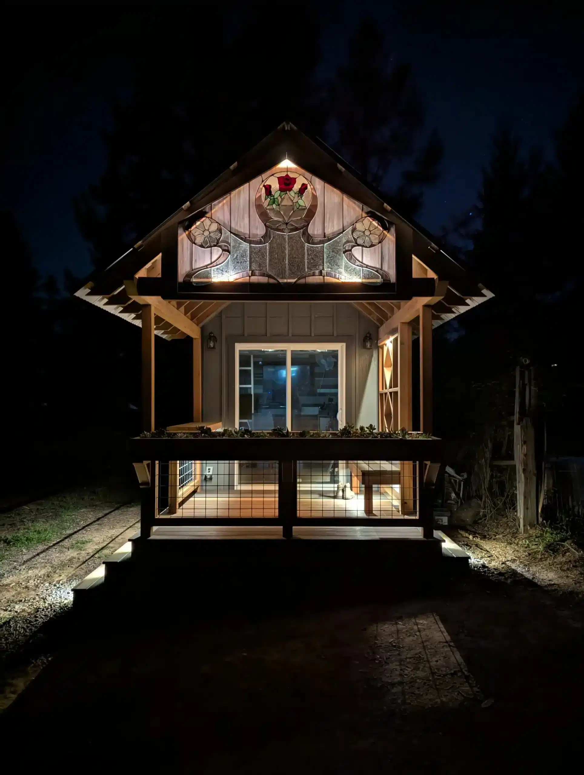 porch vs deck in napa county ca A small, illuminated wooden building at night. The building has a gable roof with a decorative stained glass window near the peak. Below the window is a covered porch area with a glass door and window. The porch is surrounded by a low wooden railing with metal mesh infill